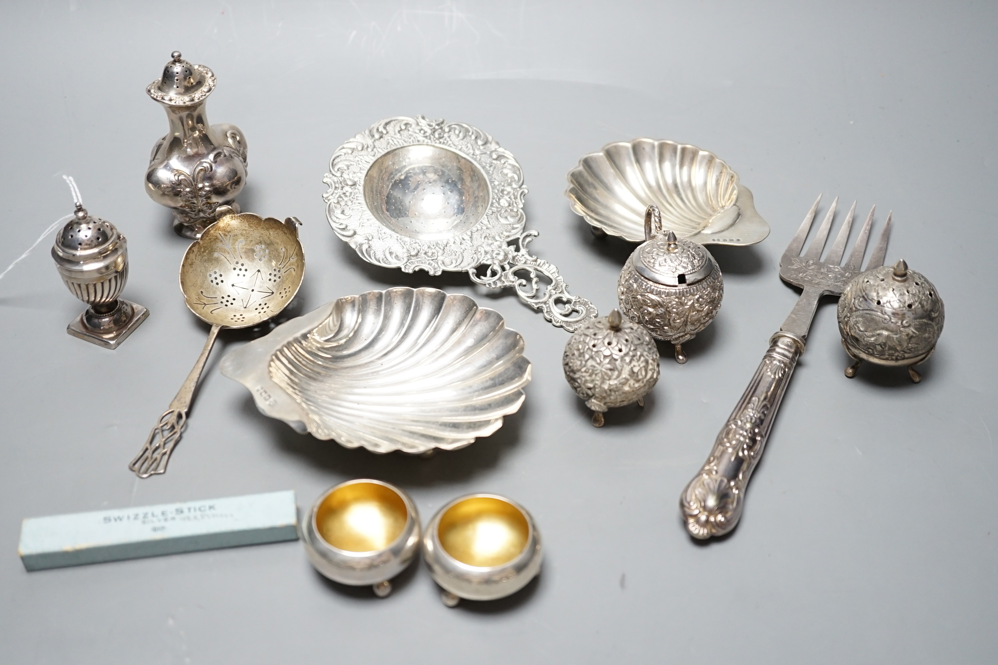 Small silver including two butter shells, a continental silver tea strainer, English tea strainer, pair of salts, swizzle stick, serving fork, two pepperettes and an Indian white metal three piece condiment set.